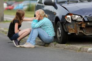 Mother and child sitting on curb with damaged car in background, after a car accident.