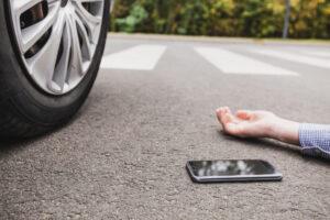 Hand and cell phone on ground with car tire in view, symbolizing a pedestrian hit by a car and the need for legal help